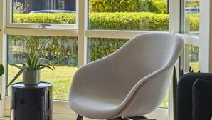 Een moderne kamer met een grijs gestoffeerde stoel, een potplant en een groot raam met uitzicht op een keurig bijgesnoeid haagje en een gebouw.