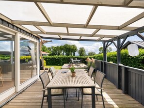 Une terrasse extérieure couverte, dotée d’une table et de chaises, une porte vitrée donnant sur un jardin et une vue sur le paysage environnant.