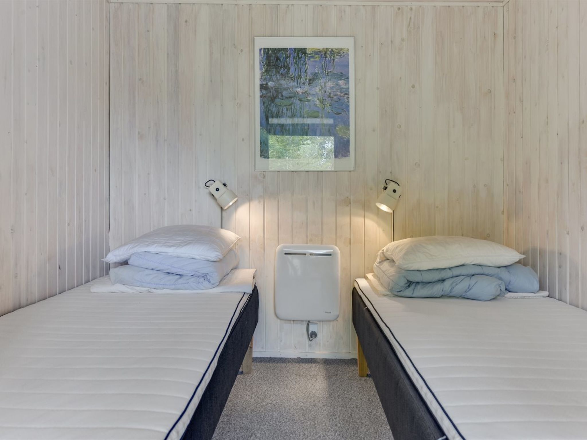 Et værelse med to senge, hver med pude og en sengebordlampe. Der er en radiator mellem sengene og et indrammet billede på væggen.