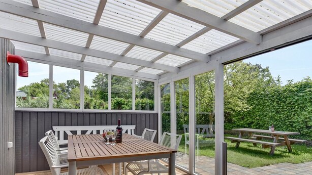 Een overdekt terras met een houten tafel en stoelen, een fles wijn en uitzicht op een tuin.