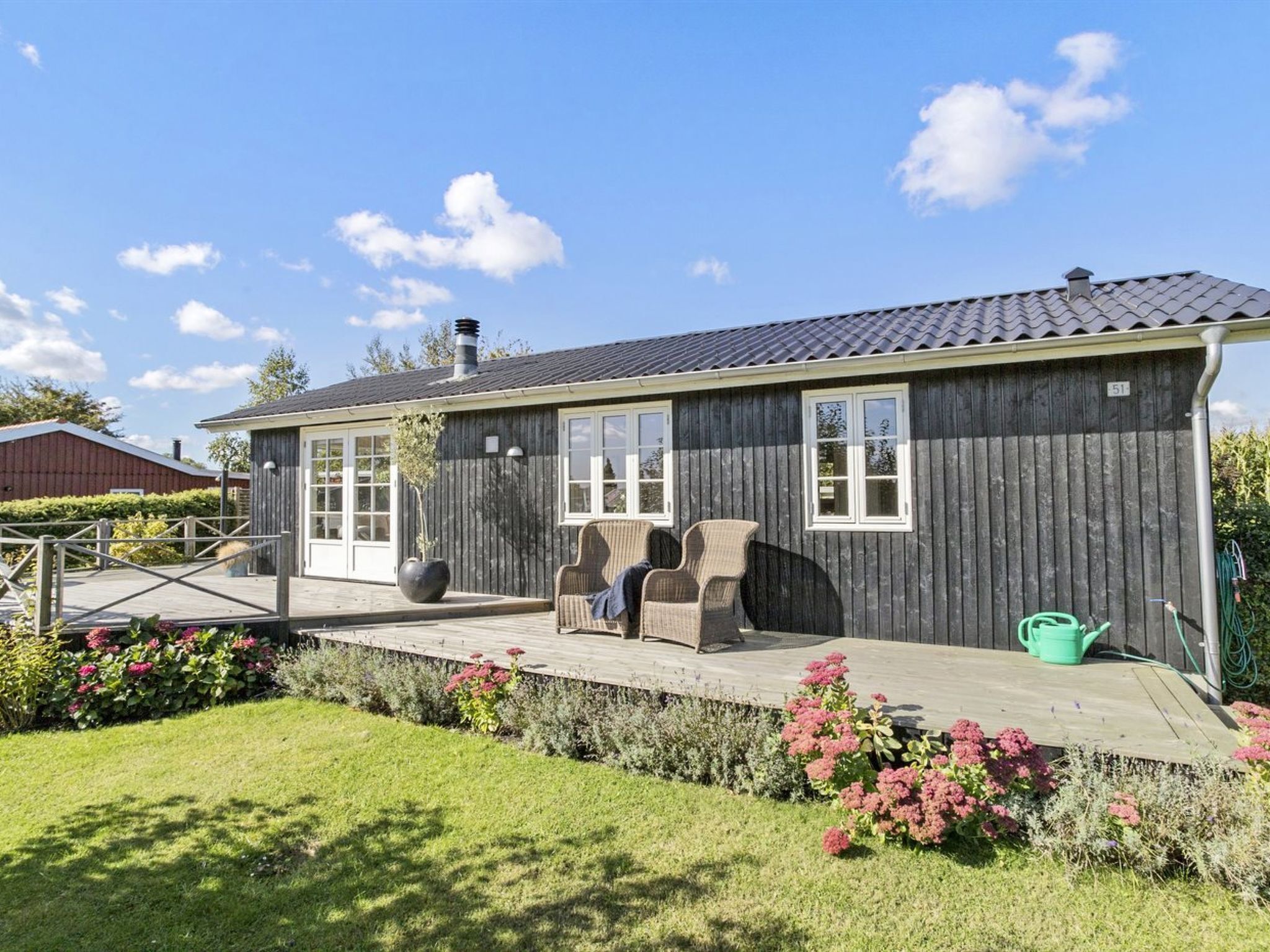 Ein Holzhaus mit überdachter Terrasse, Sitzgelegenheit im Freien und einem Garten mit rosa Blumen.