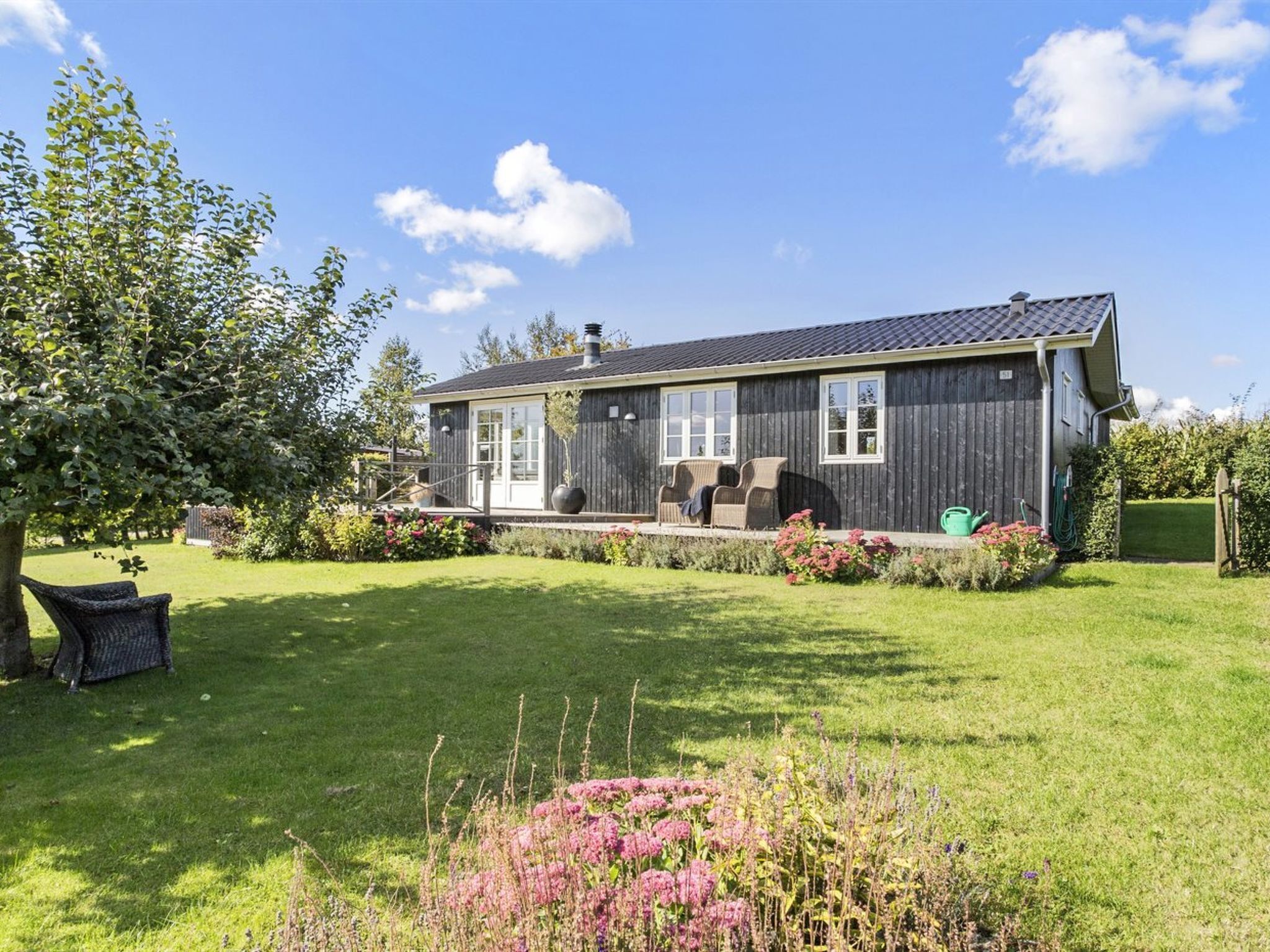 Une maison à deux étages, à l’extérieur sombre, dotée d’un porche abrité avec des meubles d’extérieur et d’une pelouse bien entretenue agrémentée de plantes à fleurs.