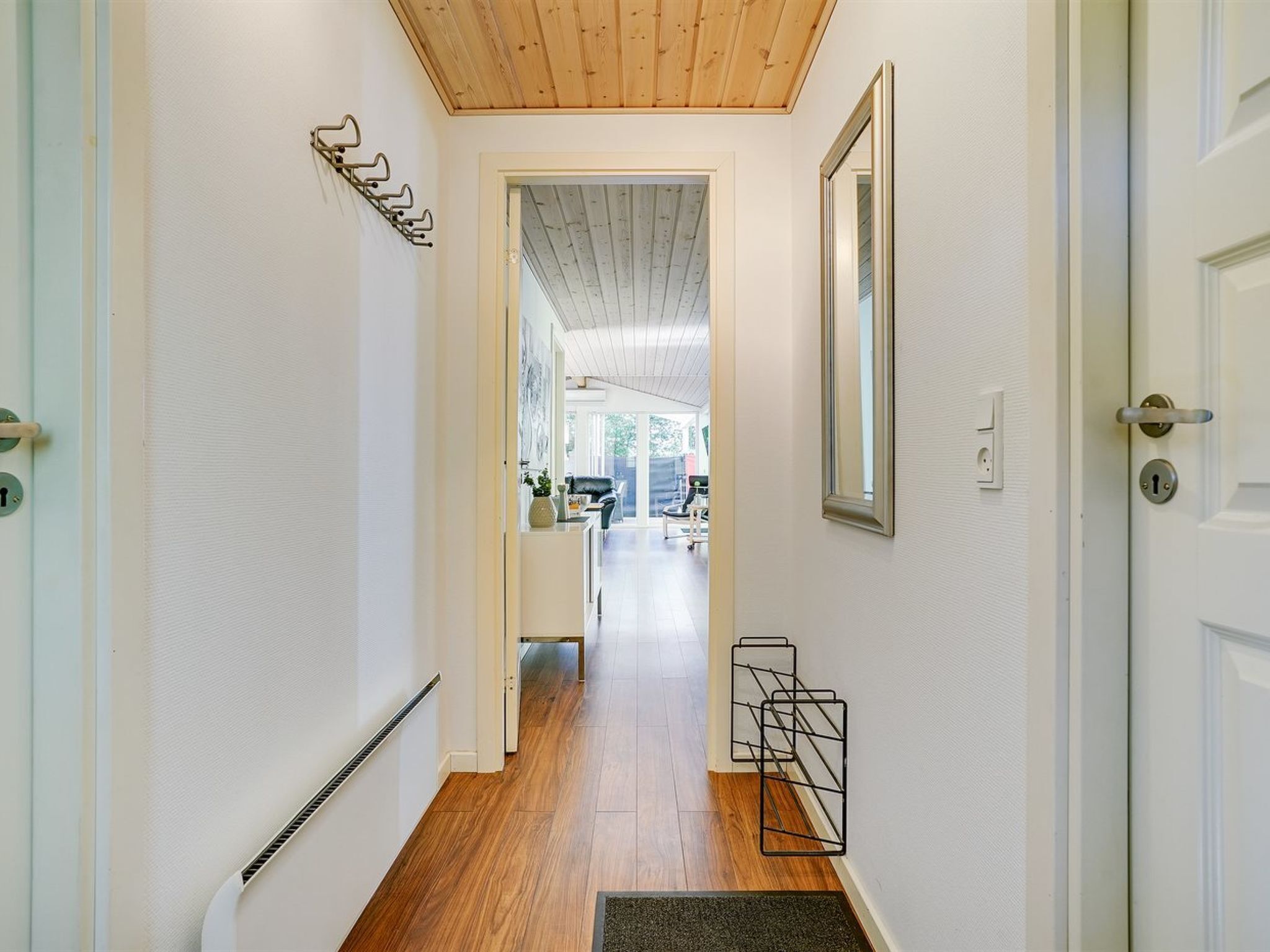 Un pasillo con suelo de madera, paredes blancas y techo con vigas de madera. Se observa una puerta a la derecha con una cerradura, y un radiador a la izquierda.