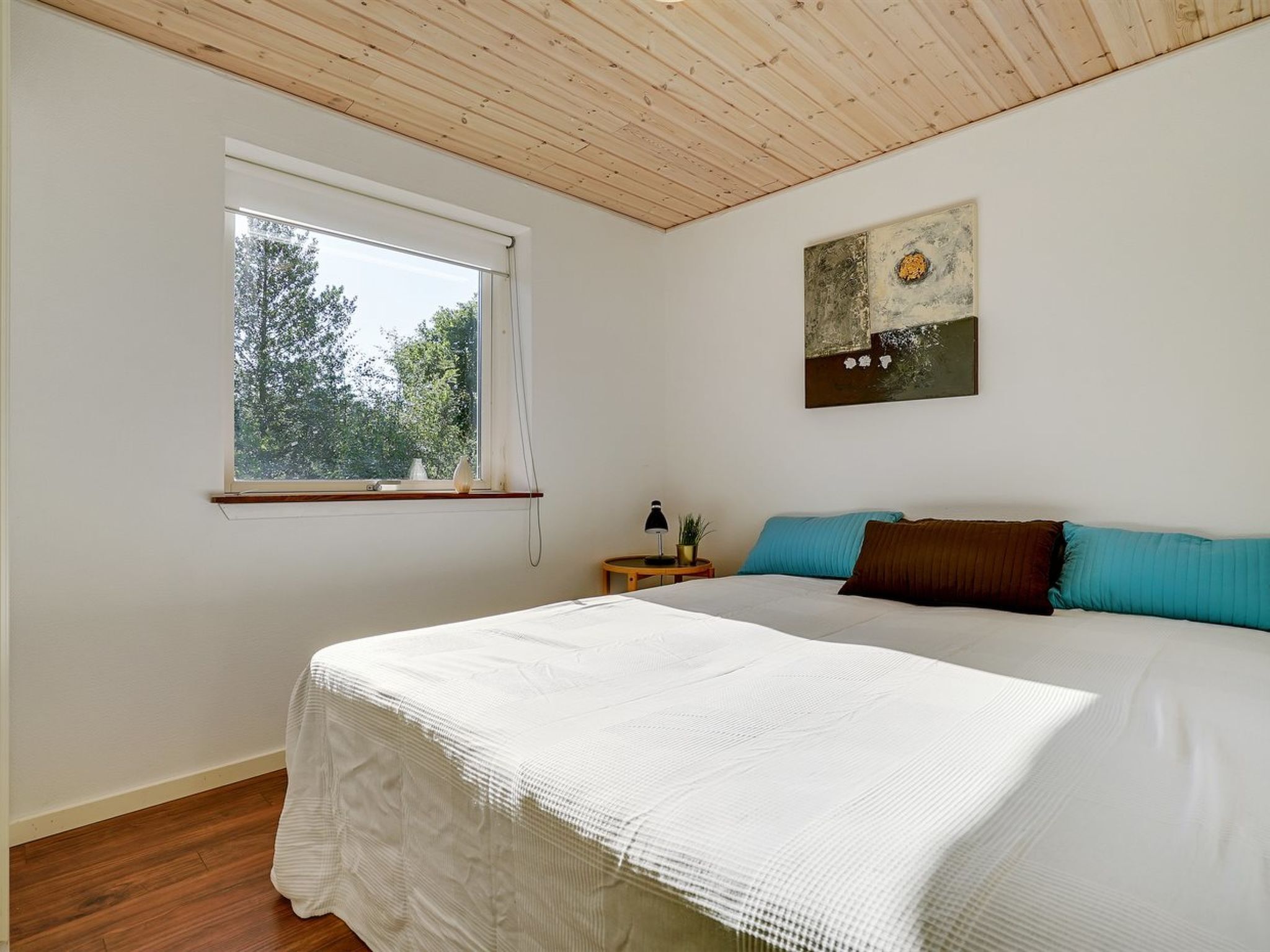 A bedroom with a wooden ceiling, a large window, a bed with white bedding, and a painting on the wall.