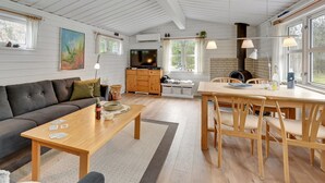 En hyggelig stue med sofa, sofabord og spiseområde med stole. Der er et fjernsyn, et tekøkken og store vinduer, der giver naturligt lys.