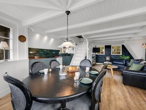 A dining area with a round table, chairs, and a chandelier. There is a kitchen in the background with a green backsplash.