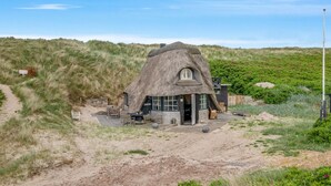 Una casa con techo de paja, visible entrada, mobiliario exterior y un entorno arenoso.
