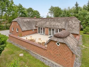 Une maison à toit de chaume, aux murs en briques, dotée d’un patio avec des meubles d’extérieur et d’une grande fenêtre.