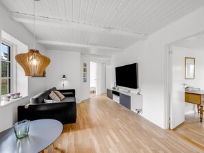 A modern living room with a black sofa, a glass coffee table, a wooden ceiling, and a flat-screen TV mounted on the wall.