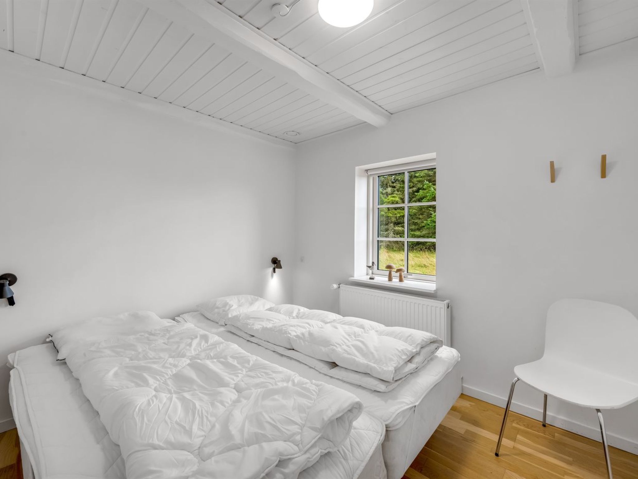 A minimalist bedroom with a large bed, a window with a view of greenery, and a white chair.