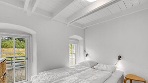Un dormitorio con una cama grande, un taburete pequeño, una ventana con vistas a zonas verdes y un techo con vigas a la vista.