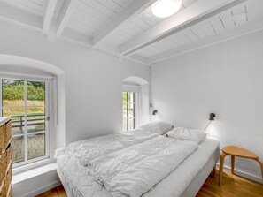 A bedroom with a large bed, a small stool, a window with a view of greenery, and a ceiling with exposed beams. - "Cornelius" - 900m from the sea by Interhome (Ulfborg)