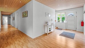 Um corredor com piso de madeira, paredes brancas e teto com vigas de madeira. Há uma porta com janela, uma pequena mesa com abajur e um extintor de incêndio na parede.