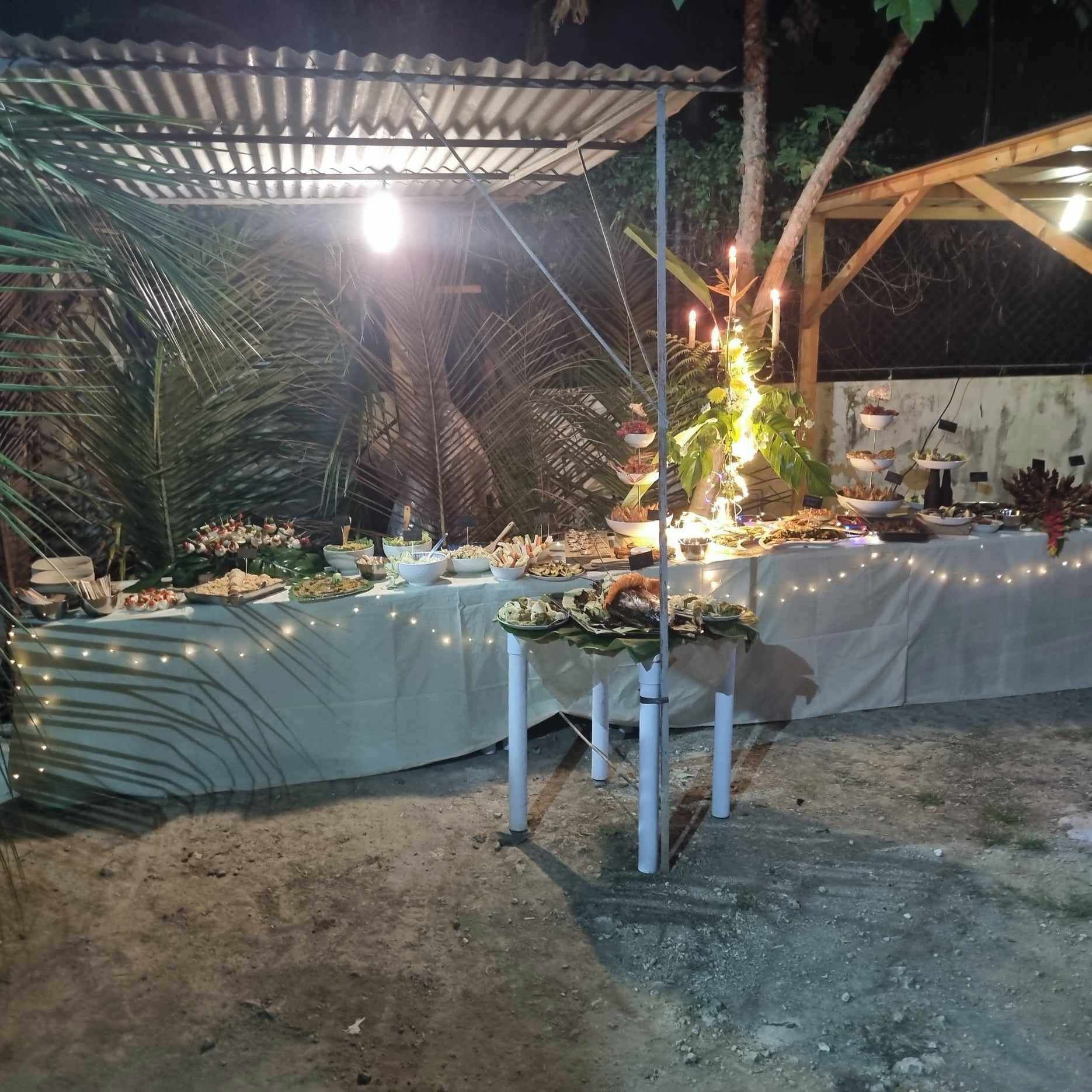 An outdoor dining area with a covered table set with various dishes, surrounded by palm trees and a metal roof structure.