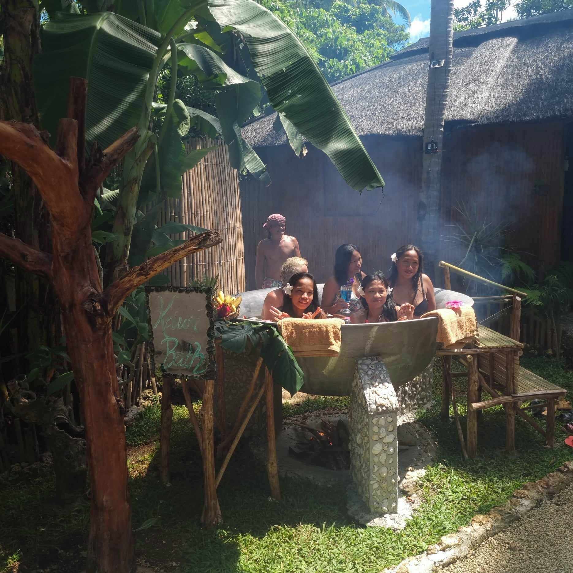 A group of people in a rustic outdoor setting with a thatched-roof structure and a wooden tub filled with water.