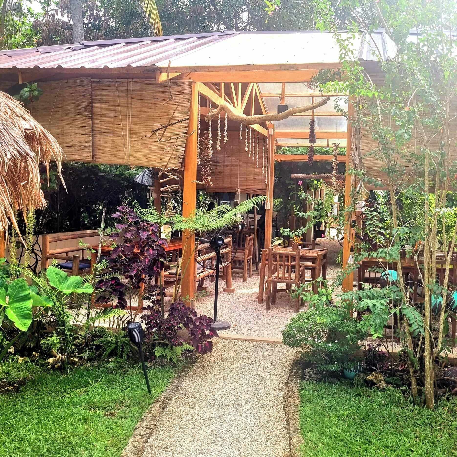 A rustic outdoor seating area with wooden tables and chairs, surrounded by lush greenery and a gravel path.