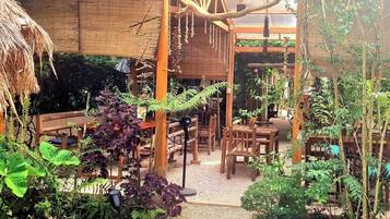A rustic outdoor seating area with wooden tables and chairs, surrounded by lush greenery and a gravel path.