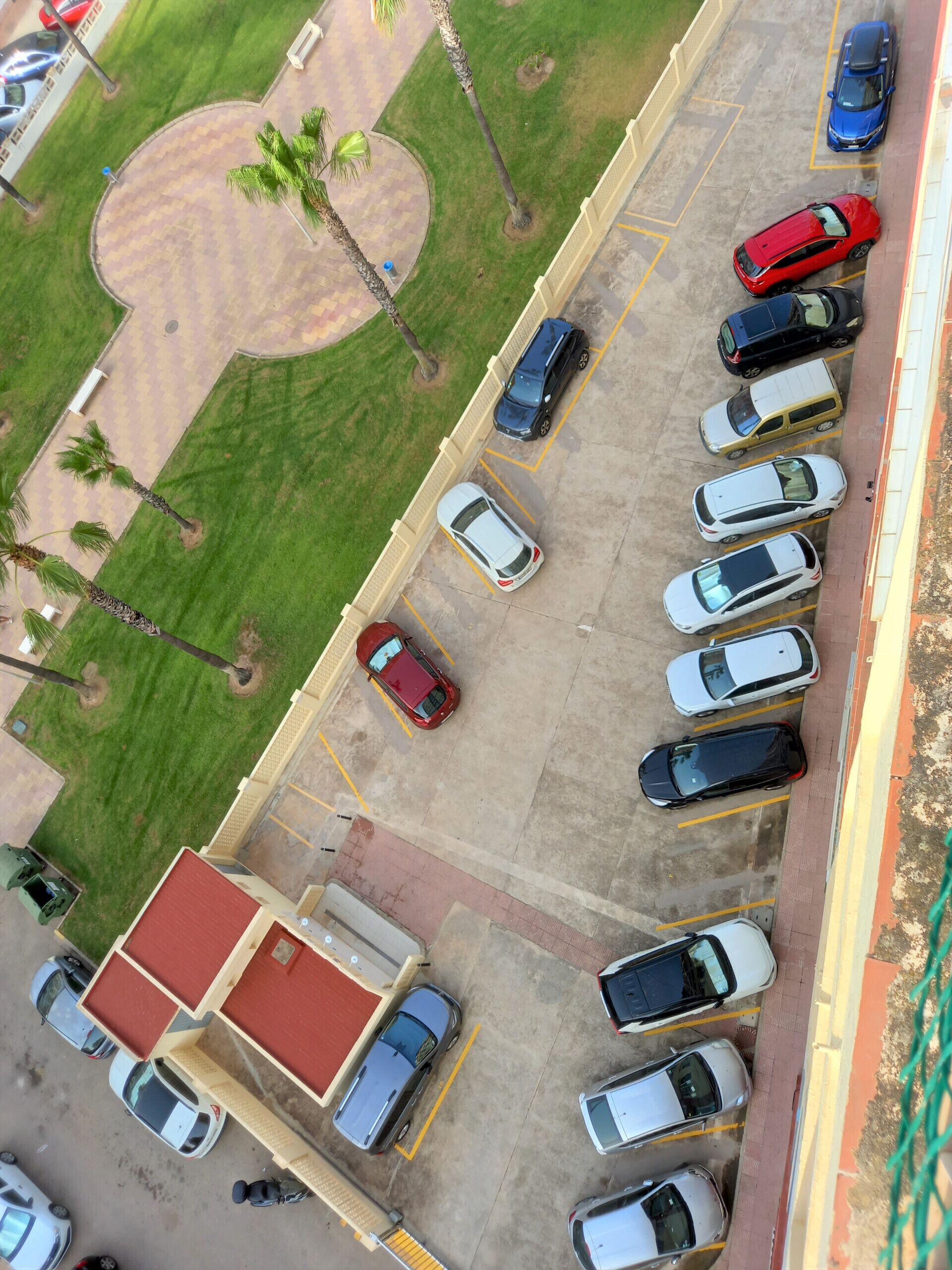 An aerial view of a parking lot with multiple cars, a building with a red roof, and palm trees.