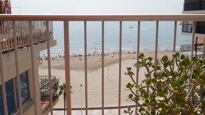 A balcony with a view of a beach, a palm tree, and a building with balconies.