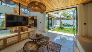A modern living room with a large flat-screen TV, wooden furniture, and a glass door leading to a garden. - Villa Arunika - 2BR Villa (Tabanan)