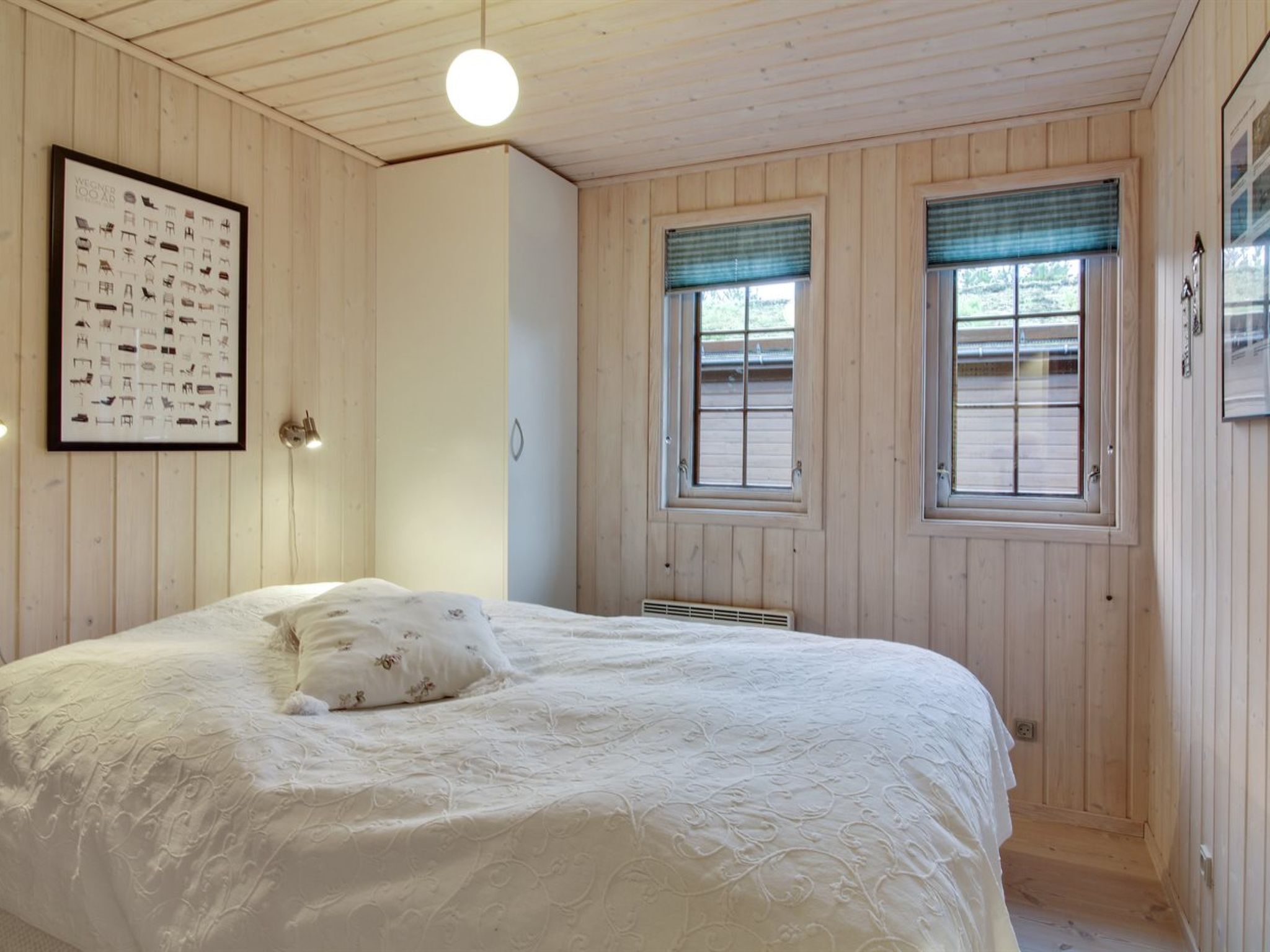 Une chambre à coucher avec un grand lit, des murs en bois, deux fenêtres équipées de stores et un tableau encadré accroché au mur.