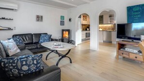 Ein modernes Wohnzimmer mit Ledersofa, Couchtisch und TV-Möbel. Es gibt einen Kamin, eine Küche mit Durchgang und eine wandmontierte Klimaanlage.