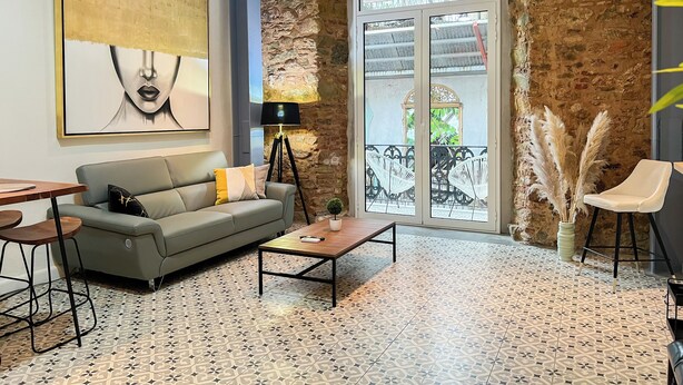 A modern living room with a grey sofa, a coffee table, a floor lamp, and a large abstract painting. A balcony with a wrought iron railing and potted plants is visible through a glass door. - Apartamento en el Corazón de Casco Viejo Panamá 14 (Panamá)