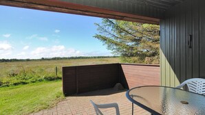 Een terras met een glazen tafel en stoelen, een houten bankje en uitzicht op een grasveld en bomen.