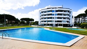 A rectangular swimming pool with a wooden deck, surrounded by a well-maintained lawn and a multi-story building with balconies. - T1 TENIS GOLFMAR - PISCINA- WIFI- VILAMOURA (Vilamoura)