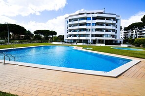 A rectangular swimming pool with a wooden deck, surrounded by a well-maintained lawn and a multi-story building with balconies. - T1 TENIS GOLFMAR - PISCINA- WIFI- VILAMOURA (Vilamoura)
