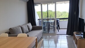 A modern living room with a sofa, dining table, and chairs. There is a balcony with a table and chairs, and a TV on a stand. - T1 TENIS GOLFMAR - PISCINA- WIFI- VILAMOURA (Vilamoura)