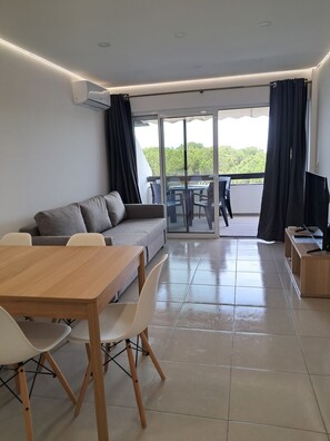 A modern living room with a sofa, dining table, and chairs. There is a balcony with a table and chairs, and a TV on a stand. - T1 TENIS GOLFMAR - PISCINA- WIFI- VILAMOURA (Vilamoura)