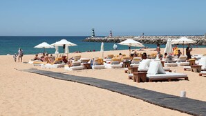 A beach with sun loungers and umbrellas, a wooden walkway, and a rocky breakwater with lighthouses in the background.
