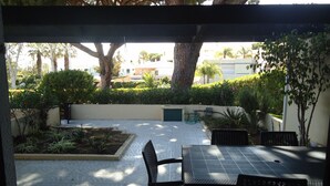 A covered patio area with a tiled floor, a table, and chairs. There are trees and a hedge in the background.