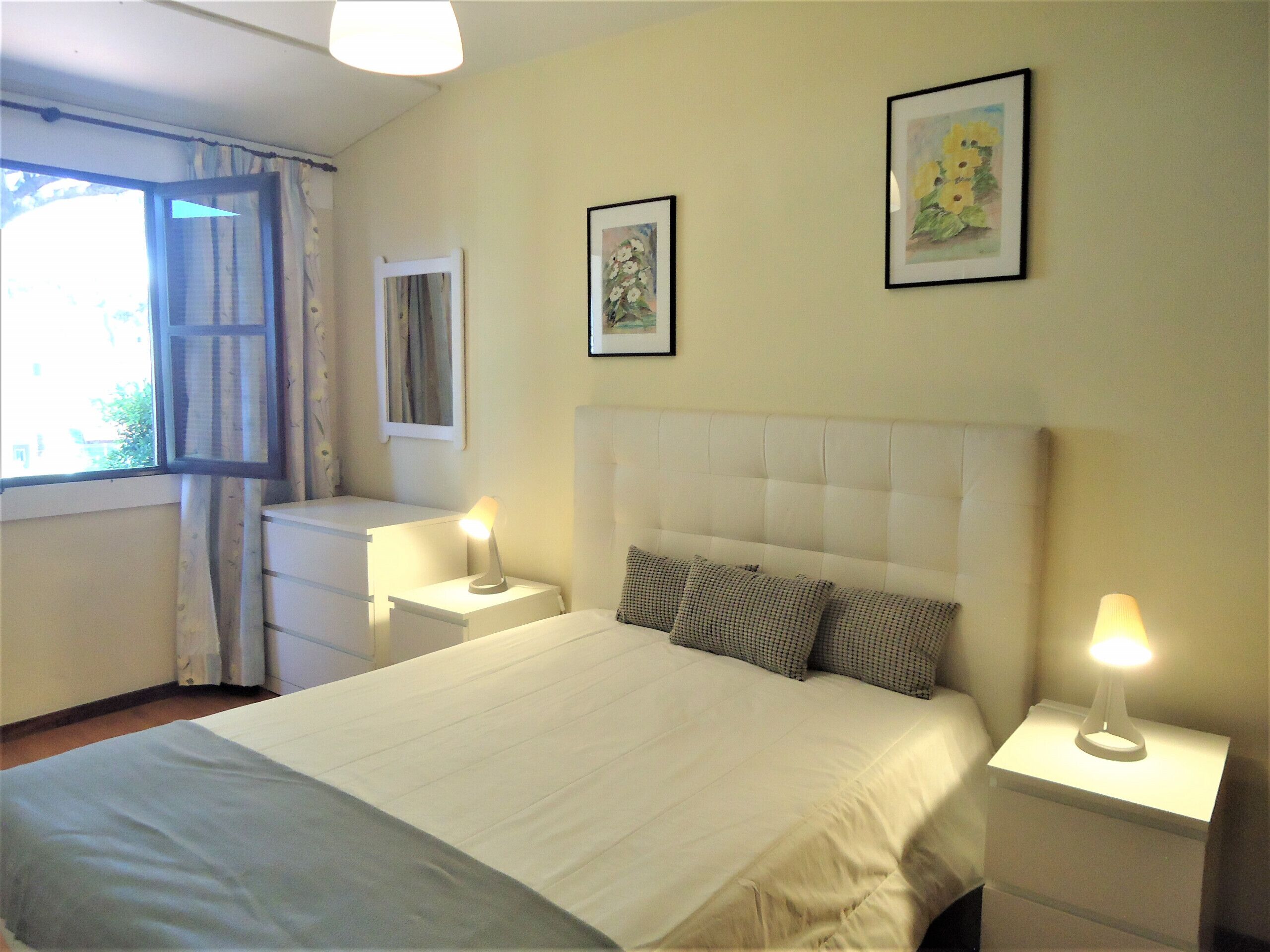 A bedroom with a bed, two bedside tables with lamps, a window with curtains, and two framed pictures on the wall.