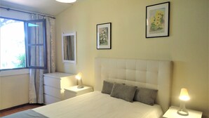 A bedroom with a bed, two bedside tables with lamps, a window with curtains, and two framed pictures on the wall.