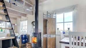 A cozy room with a wood-burning stove, a white chair, a dining table, and a window with curtains.