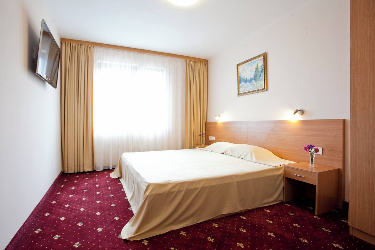 A hotel room with a bed, a bedside table, a nightstand, a lamp, a painting, and a window with curtains.