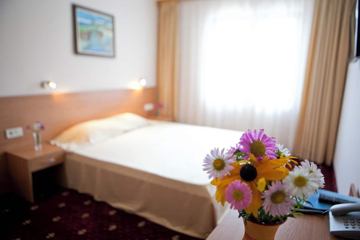 A hotel room with a bed, a nightstand, a vase with flowers, and a window with curtains.