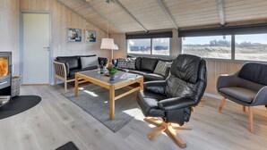 Ein gemütliches Wohnzimmer mit Kamin, Ledersofas, einem Holztisch und großen Fenstern mit Blick ins Freie.