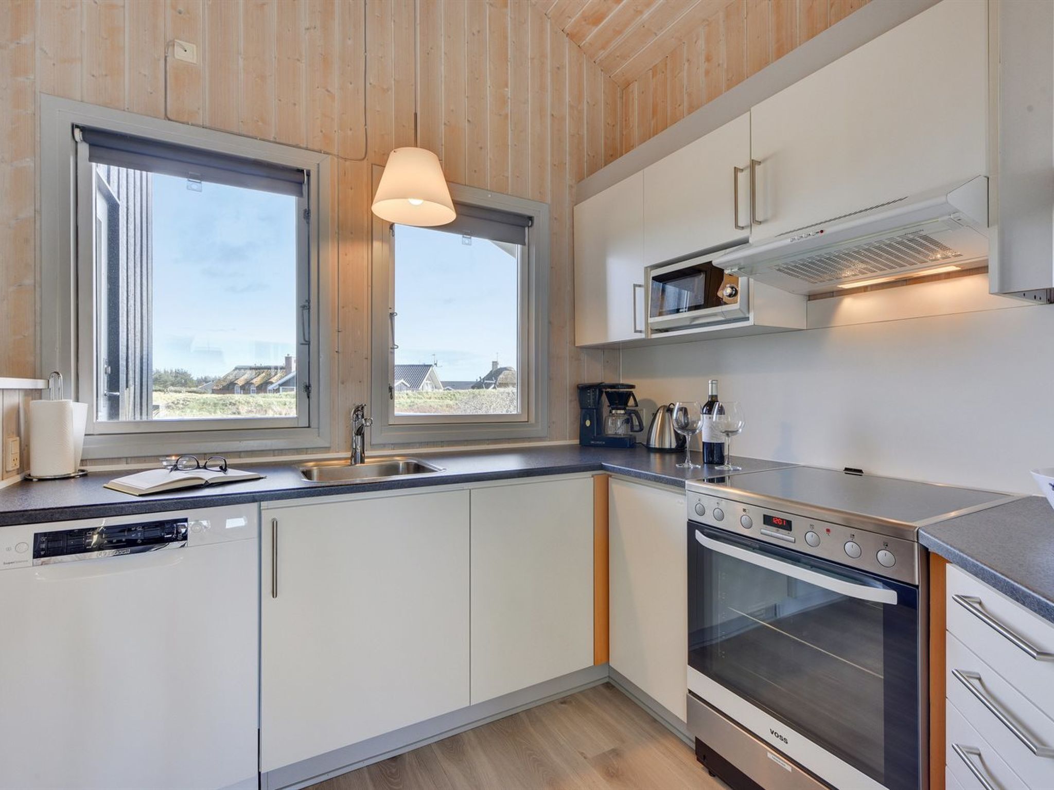 Eine moderne Küche mit weißen Schränken, einem Einbauofen und einer Mikrowelle. Zwei Fenster sorgen für natürliches Licht und bieten einen Ausblick nach draußen.