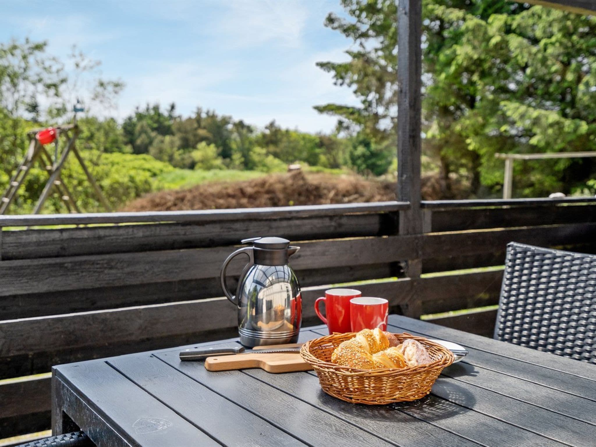 Une table en bois, une cafetière en métal, des tasses rouges, un panier en osier contenant du pain et une planche à découper en bois.