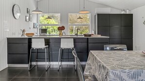Une cuisine moderne dotée d’un îlot central, de deux tabourets de bar, d’un éclairage suspendu et offrant une vue sur la verdure à l’extérieur.
