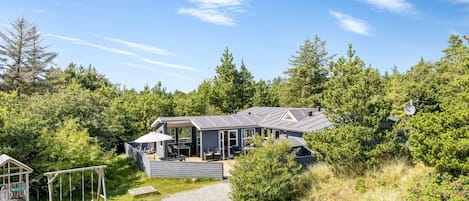 Et moderne hus med en overdækket terrasse, omgivet af tæt skov og en grussti.