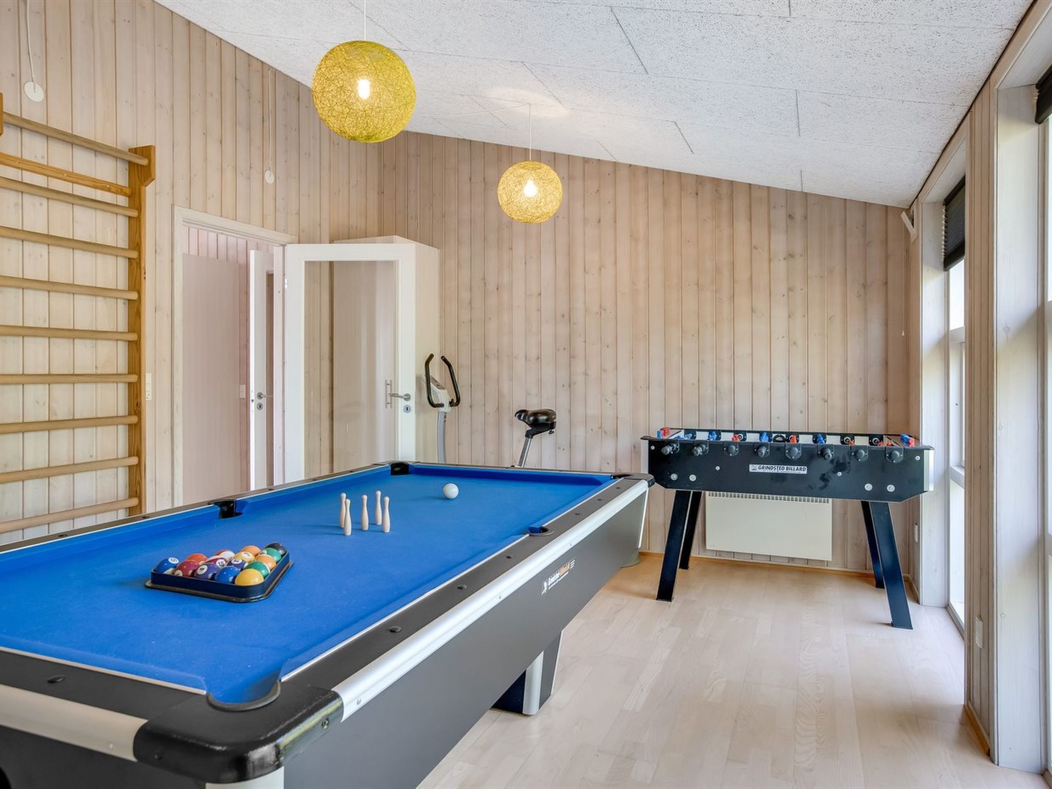 Una mesa de billar con superficie de fieltro azul y una mesa de futbolín en una habitación con paredes de madera y techo blanco.