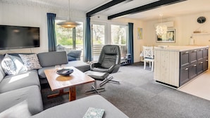 Una sala de estar moderna con un sofá, una mesa de centro y un televisor. Cuenta con una zona de comedor con una mesa y sillas. La habitación tiene ventanas amplias y una cocina con armarios.