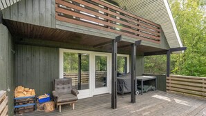 Una terraza de madera con un área de estar techada, una parrilla y una pila de leña.