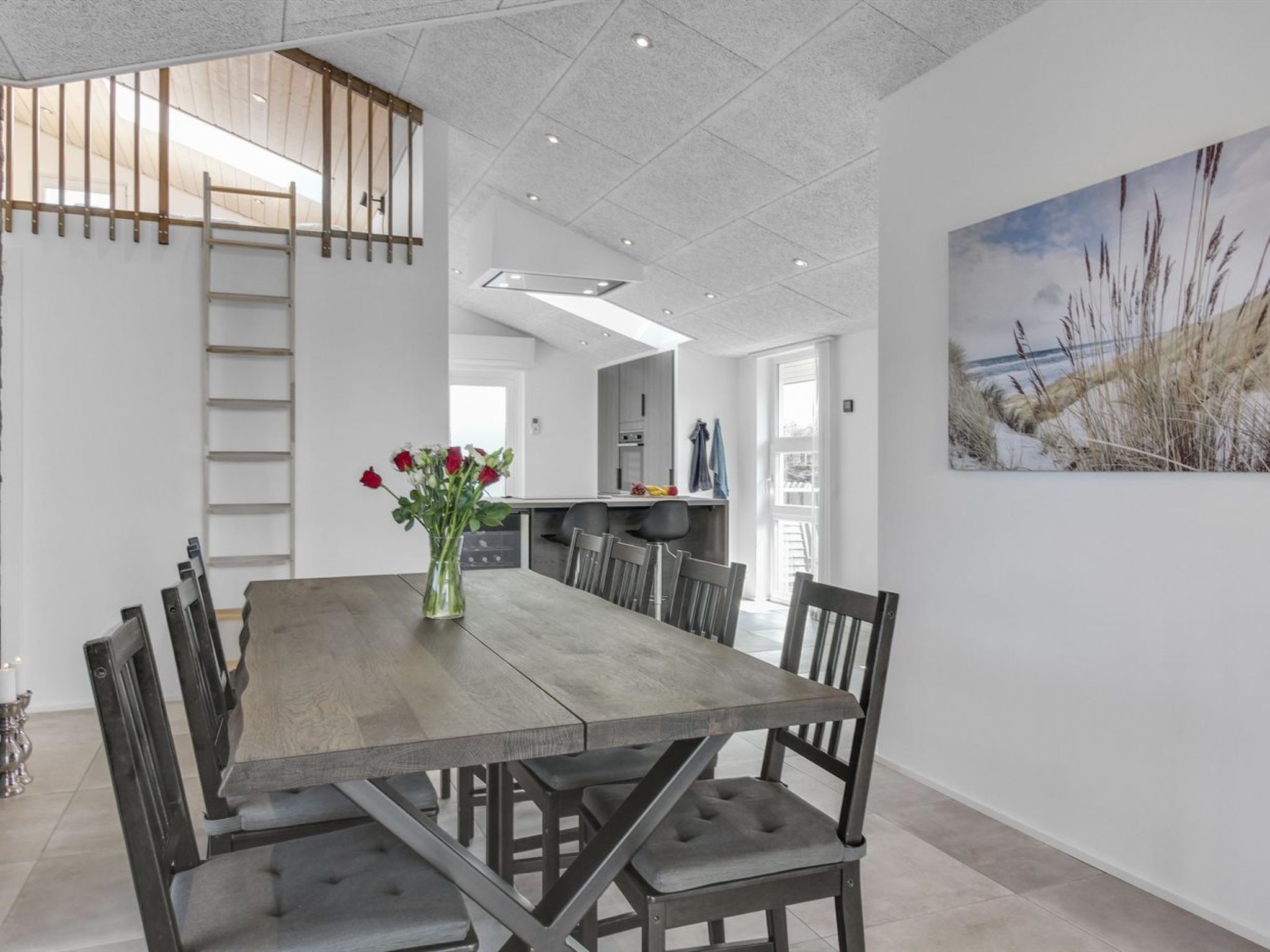 A modern dining area with a wooden table and chairs, a vase with flowers, and a painting of a beach scene on the wall.