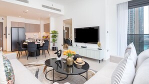 Una sala de estar moderna con un sofá, un comedor con sillas, un televisor montado en la pared y un ventanal con vistas a la ciudad.