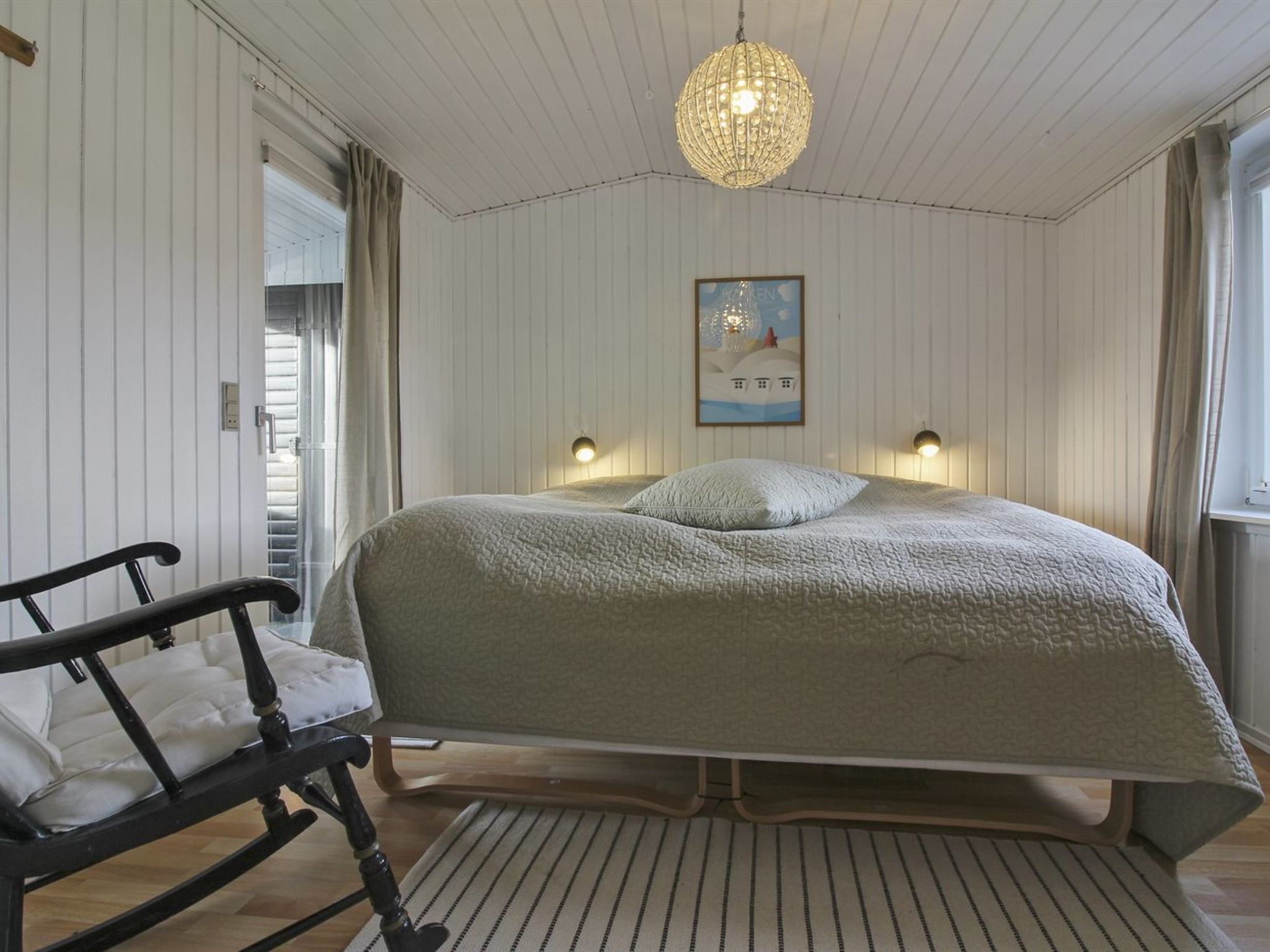 A bedroom with a bed, a chair, a rug, a chandelier, and a painting on the wall.